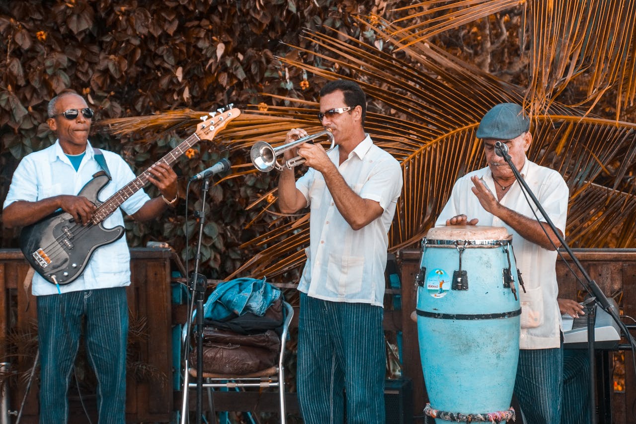 Three musicians perform with trumpet, bass guitar, and conga in an outdoor setting.